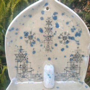 Puede incluir: Un porta incienso de cerámica color crema con motas azules y diseños geométricos. El soporte tiene un pequeño soporte cilíndrico para varillas de incienso y un agujero en la parte superior. El diseño incluye patrones de copos de nieve y estrellas.