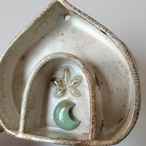 May include: A white ceramic wall hanging with a crescent moon and a stylised flower design. The hanging has three compartments.