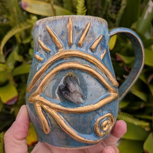 Puede incluir: Una taza de cerámica azul con un diseño de ojo en relieve de color dorado, con rayos de sol arriba y una espiral abajo. Un pequeño cristal oscuro e irregular está incrustado en el centro del ojo. La taza cuenta con un asa curva y esmalte.