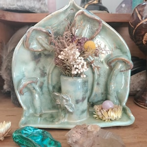May include: A teal ceramic decorative piece with mushroom and leaf designs, holding dried flowers. A small vase is in the center. A malachite stone, a quartz crystal, and a small nest with a pink stone are in front.