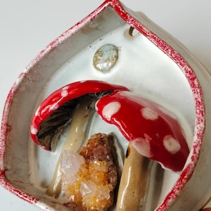 May include: A white ceramic planter shaped like a house with a red rim. The planter features two red and white mushrooms with brown stems and a clear quartz crystal.