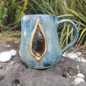 May include: A blue ceramic mug with a gold-trimmed, teardrop-shaped window containing a dark crystal. The mug has a handle and a textured surface. The mug is sitting on a gray rock.