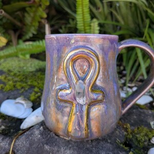 Könnte beinhalten: Ein Keramikbecher mit glänzender, irisierender Oberfläche in Violett- und Kupfertönen. Auf der Vorderseite befindet sich ein erhabenes, gold bemaltes Ankh-Symbol mit einem kleinen, klaren, spitzen Kristall in der Mitte und einem geschwungenen Henkel.