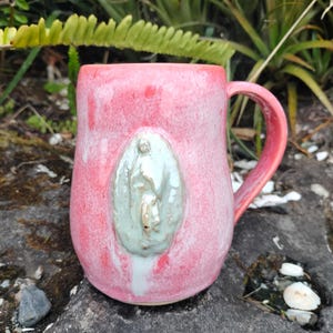 May include: A pink glazed ceramic mug with a light green religious figure relief. The mug has a rounded shape with a handle. The mug is set against a natural background of rocks and greenery.