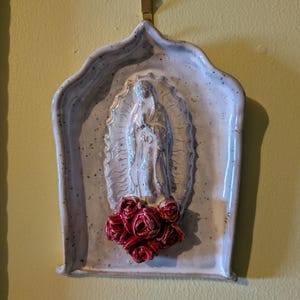 Puede incluir: Una placa de cerámica para pared con un marco arqueado de color gris claro moteado, que presenta un relieve central de la Virgen de Guadalupe. La figura está rodeada por un halo radiante y adornada con rosas rojas esculpidas en la base.