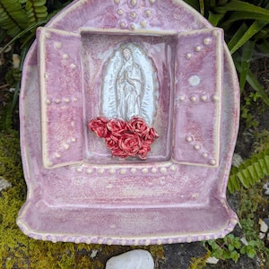 Puede incluir: Un santuario de pared de cerámica color lavanda con una parte superior arqueada y dos puertas abiertas. Una figura en relieve blanca de la Virgen de Guadalupe se encuentra en el centro, rodeada por un grupo de rosas rojas esculpidas en la base.