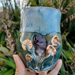 Op de afbeelding: Een handgemaakte keramische mok met een turquoise en groene glazuur. De mok heeft goudkleurige paddenstoelaccenten en een centrale cluster van kristallen. De mok wordt in een hand gehouden, wat het unieke ontwerp laat zien.