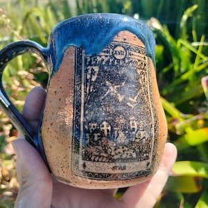 May include: Handmade ceramic mug with a blue glazed rim and handle. The mug features a detailed black and white illustration of a tarot card. The mug has a tan body with a textured surface, perfect for tea or coffee.