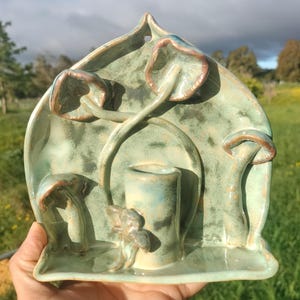 Pode incluir: Uma escultura em cerâmica em tons de verde e castanho, com um design estilizado de cogumelos. A peça tem um elemento cilíndrico central e um fundo curvo em forma de folha. A escultura é segurada na mão contra um fundo exterior desfocado.