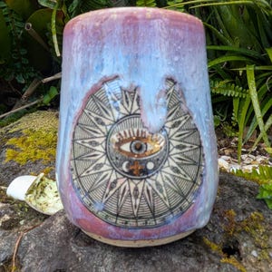 May include: A ceramic tumbler with a glossy, multi-coloured glaze in shades of pink, lavender, and white. The front features a detailed black and gold circular graphic of an eye surrounded by radiating lines, sun rays, and celestial symbols.