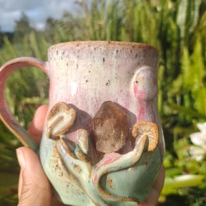 May include: A handcrafted ceramic mug with a light pink and green glaze. The mug features a sculpted design with a brown crystal and mushroom accents. The handle and rim have a speckled finish. A unique and artistic piece for enjoying beverages.