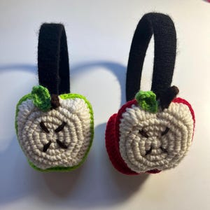 Op de afbeelding: Een paar gebreide appel oorwarmers. De ene oorwarmer is rood en de andere is groen. Beide oorwarmers hebben een wit gebreid appelontwerp met een bruine steel en een groen blad.