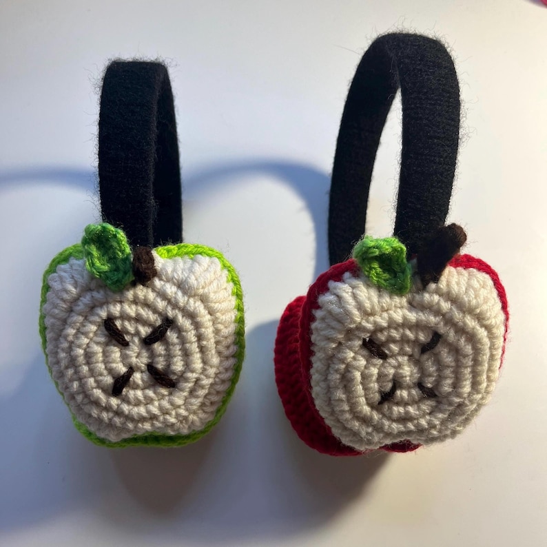 Op de afbeelding: Een paar gebreide appel oorwarmers. De ene oorwarmer is rood en de andere is groen. Beide oorwarmers hebben een wit gebreid appelontwerp met een bruine steel en een groen blad.