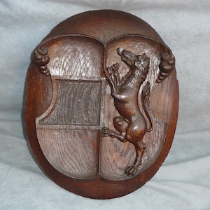 Könnte beinhalten: Ovale Holz-Wandplakette mit geschnitztem Design. Die Plakette hat eine Schildform mit einer detaillierten Reliefschnitzerei eines Löwen. Das Holz hat einen warmen, braunen Ton und die Schnitzerei ist aufwendig.