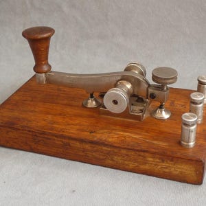 Op de afbeelding: Een antieke telegraafsleutel, met een houten basis en een metalen mechanisme. De sleutel heeft een houten handvat en verschillende metalen knoppen en palen. Het hout heeft een rijke, warme tint en de metalen componenten hebben een zilveren afwerking. Een vintage communicatieapparaat.