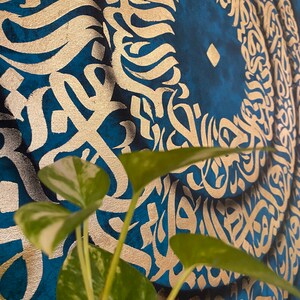 Puede incluir: Un primer plano de una pintura abstracta azul y dorada con un dise&ntilde;o circular. La pintura presenta intrincados patrones y remolinos, con un c&iacute;rculo central que parece ser un ojo estilizado. Una planta verde est&aacute; en primer plano.