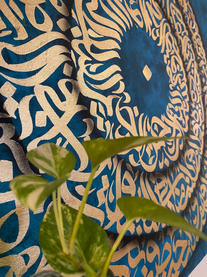 Puede incluir: Un primer plano de una pintura abstracta azul y dorada con un dise&ntilde;o circular. La pintura presenta intrincados patrones y remolinos, con un c&iacute;rculo central que parece ser un ojo estilizado. Una planta verde est&aacute; en primer plano.