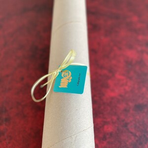 Puede incluir: Un p&oacute;ster o impresi&oacute;n enrollado con una cinta amarilla atada alrededor. Una etiqueta azul con el texto "Calligraphy Art" est&aacute; unida a la cinta.