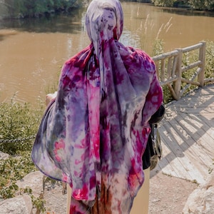 May include: A person wearing a tie-dye hijab in shades of purple, pink, and gray, standing near a wooden bridge and a body of water. The scarf is draped over the head and shoulders, creating a flowing, colorful effect.