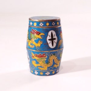 May include: A small, blue, cylindrical container with a lid, decorated with gold and yellow dragons and a black star. The container is made of metal and has a glossy finish.