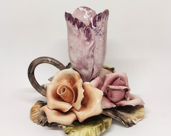 Op de afbeelding: Een keramische kaarsenhouder met een roze tulp en twee rozen. De rozen zijn in tinten roze en oranje. De kaarsenhouder is versierd met groene bladeren.