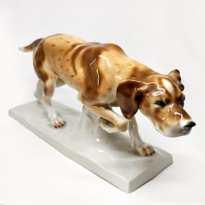 Puede incluir: Una figurita de porcelana de un perro de caza marrón y blanco en una pose de carrera. El perro se representa con la cabeza girada hacia un lado, como si estuviera siguiendo un olor. La figurita está colocada sobre una base rectangular blanca.