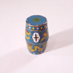 May include: A small, blue, cylindrical container with a lid. The container is decorated with a gold trim and intricate designs of dragons, birds, and clouds. The lid has a green flower design.
