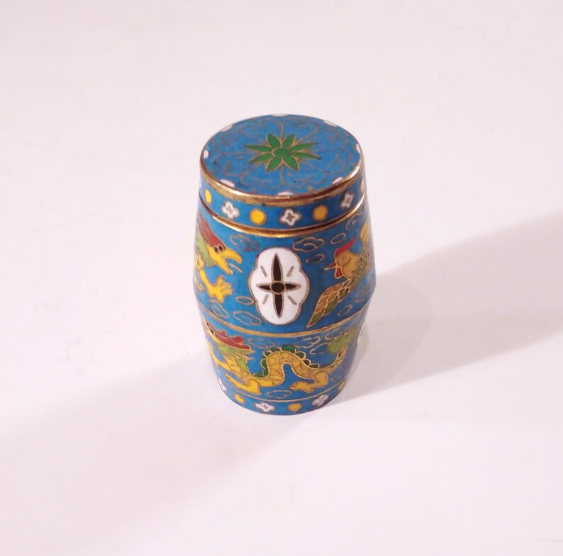 May include: A small, blue, cylindrical container with a lid. The container is decorated with a gold trim and intricate designs of dragons, birds, and clouds. The lid has a green flower design.