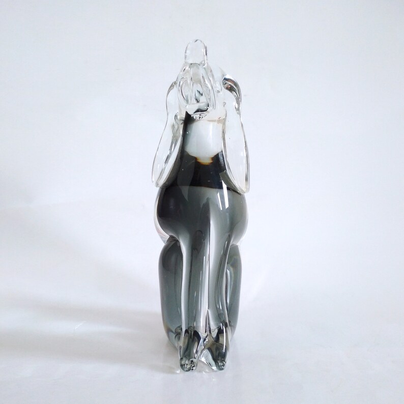 Pu&ograve; includere: Scultura in vetro trasparente e grigio raffigurante un cane seduto, con grandi orecchie. La scultura ha una finitura liscia e lucida, su sfondo bianco. Il corpo del cane &egrave; di colore grigio scuro.