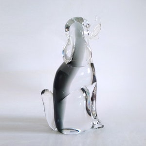 Pu&ograve; includere: Una statuetta di cane in vetro trasparente in posizione seduta. La scultura presenta una sfumatura di grigio e vetro trasparente, con grandi orecchie curve e una piccola coda. Il cane &egrave; seduto in posizione eretta, con la testa leggermente inclinata verso l'alto.