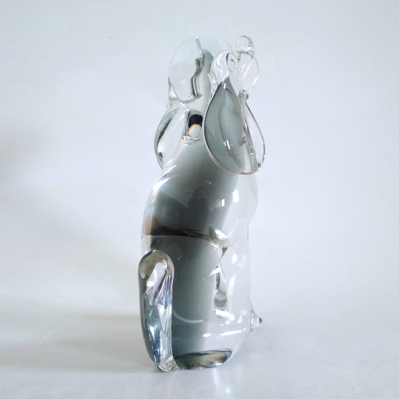 Pu&ograve; includere: Una scultura in vetro trasparente e grigio raffigurante un cane. La scultura presenta grandi orecchie flosce e una coda ricurva. Il corpo del cane &egrave; grigio pieno, mentre le orecchie e la coda sono trasparenti. La scultura &egrave; su una superficie bianca.