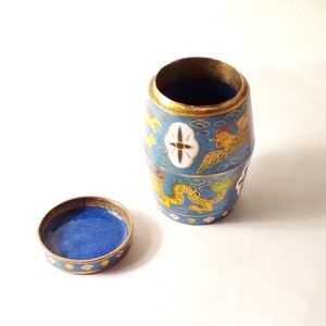 May include: A small, blue and gold cylindrical container with a lid. The container is decorated with a dragon, a bird, and clouds. The lid is also decorated with a similar design.