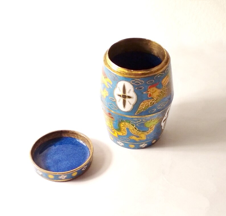 May include: A small, blue and gold cylindrical container with a lid. The container is decorated with a dragon, a bird, and clouds. The lid is also decorated with a similar design.