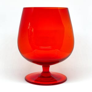 Può includere: Un grande bicchiere da brandy rosso vivo. Il bicchiere presenta una coppa arrotondata e un gambo corto con una base circolare. Il vetro trasparente riflette la luce, mettendo in risalto la sua superficie liscia e lucida. Il design è semplice ma elegante.