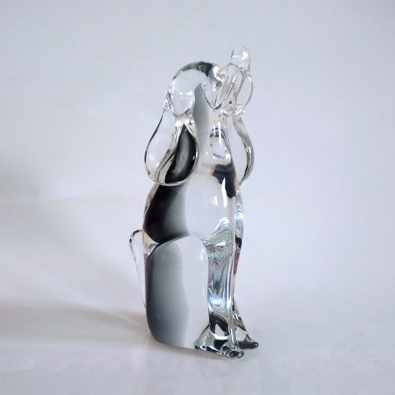 Pu&ograve; includere: Una statuetta di cane in vetro trasparente in posizione seduta, con le orecchie dritte e la coda alzata. La scultura presenta una sfumatura di nero e grigio all'interno del vetro, aggiungendo profondit&agrave;. Il cane &egrave; su sfondo bianco.