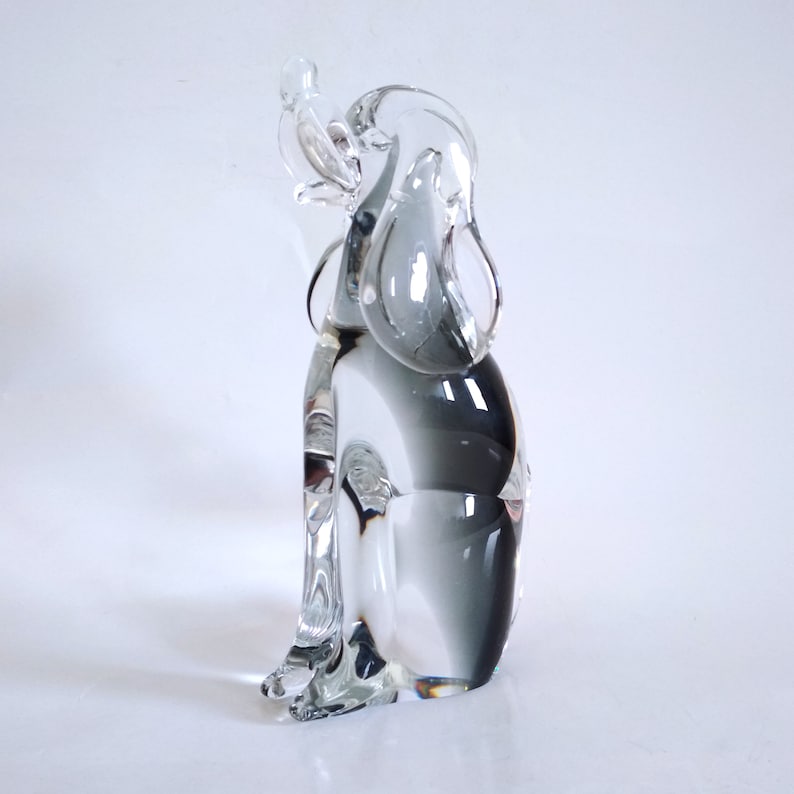 Pu&ograve; includere: Una scultura in vetro trasparente di un cane con un uccello appollaiato sulla testa. Il cane &egrave; seduto, con le orecchie abbassate. La scultura ha un effetto sfumato, che passa dal trasparente a un grigio fumoso.