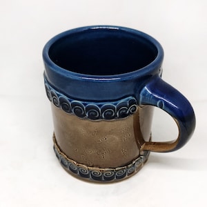 Puede incluir: Una taza de cerámica azul y marrón con un diseño decorativo alrededor del borde. La taza tiene un asa y está hecha de gres.