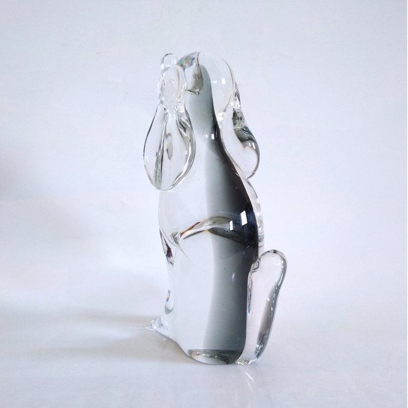 Pu&ograve; includere: Una statuetta di cane in vetro trasparente, con lunghe orecchie pendenti e una postura seduta. La scultura &egrave; trasparente, permettendo alla luce di passare attraverso, ed &egrave; posta su uno sfondo bianco. La forma del cane &egrave; liscia e dettagliata.