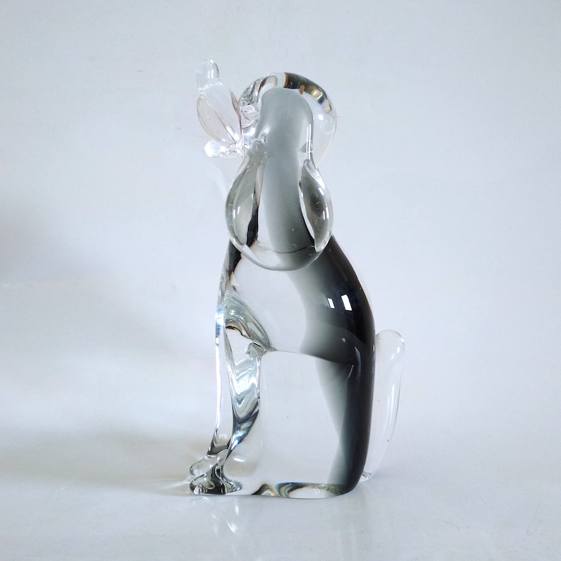 Pu&ograve; includere: Una statuetta di cane in vetro trasparente in posizione seduta. La scultura ha una finitura liscia e lucida e una sfumatura di grigio dalla base alla schiena. Le orecchie del cane sono dritte e ha un piccolo uccello trasparente appollaiato sulla testa.