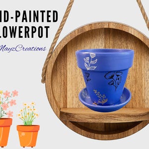 May include: Hand-painted flowerpot in a vibrant blue hue, adorned with delicate floral designs. The pot sits on a matching saucer, displayed on a wooden shelf. The image also features two orange flower pots with plants.