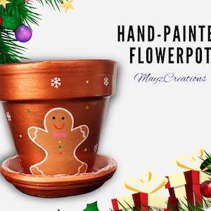 May include: Hand-painted terracotta flowerpot with a gingerbread man design. The pot is copper-colored with white snowflake accents and a matching saucer. The text "HAND-PAINTED FLOWERPOT" is displayed in black.