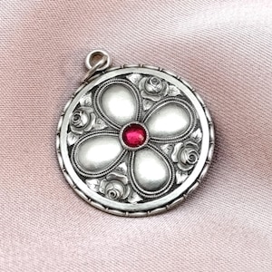 以下が含まれることがあります： 花のデザインのシルバーペンダント。ペンダントは、5つの白い花びらに囲まれた中央の赤い宝石が特徴です。外側の端は花の模様で詳細に描かれています。ペンダントは丸く、チェーン用の小さなループが上部にあります。