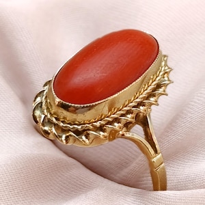 May include: A gold ring featuring an oval, vibrant red gemstone. The ring has an ornate, textured gold setting with a detailed design around the gemstone. The ring's band is a simple, polished gold.