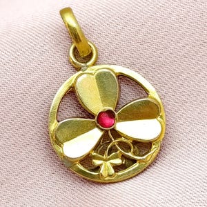 Può includere: Ciondolo dorato con design a trifoglio a tre foglie con petali a forma di cuore e una piccola gemma rossa al centro. Il ciondolo ha una cornice circolare e un anello per una catena. Lo sfondo è in tessuto rosa chiaro.
