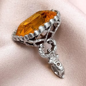 May include: A silver pendant with an oval, amber-colored gemstone. The gemstone is set in a detailed, openwork silver frame. The pendant has a decorative bail with intricate scrollwork. The background is a soft, pink fabric.