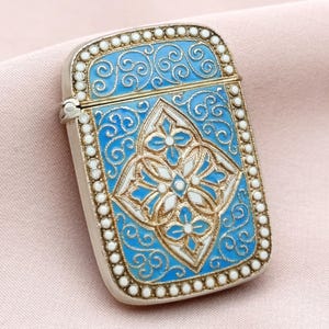 Puede incluir: Un estuche de cerillas rectangular plateado con tapa abisagrada. El estuche de cerillas presenta un diseño floral central azul y blanco, rodeado de intrincados arabescos y un borde de pequeñas cuentas blancas. El fondo es rosa pálido.