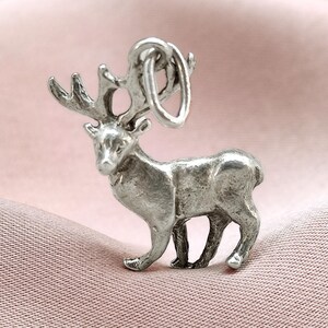 May include: A silver-colored charm in the shape of a deer with large antlers. The charm is detailed with realistic features and has a loop at the top for attaching to a bracelet or necklace. The charm is set against a pink background.