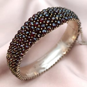 Può includere: Un bracciale vintage a cerniera con montatura in metallo argentato. Il bracciale è incrostato di numerose piccole gemme rotonde, rosso scuro. Le gemme sono ravvicinate, creando una superficie continua e strutturata. Il bracciale è esposto su un tessuto rosa.