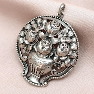 May include: A silver pendant featuring a floral design. The pendant showcases a vase overflowing with roses and other flowers, surrounded by a decorative border. The pendant has a loop at the top for a chain.