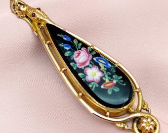May include: A gold-toned pendant with a teardrop-shaped black enamel center. The enamel features a floral design with pink, blue, and white flowers. The pendant has a snake-like detail at the top and a floral accent at the bottom.